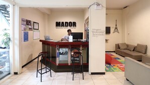 Una recepción con un hombre trabajando, un cartel que dice "MADOR MALANG DORM HOSTEL" y un letrero de bienvenida con la inscripción "Welcome! Bienvenue! Benvenuto! Willkommen!".
