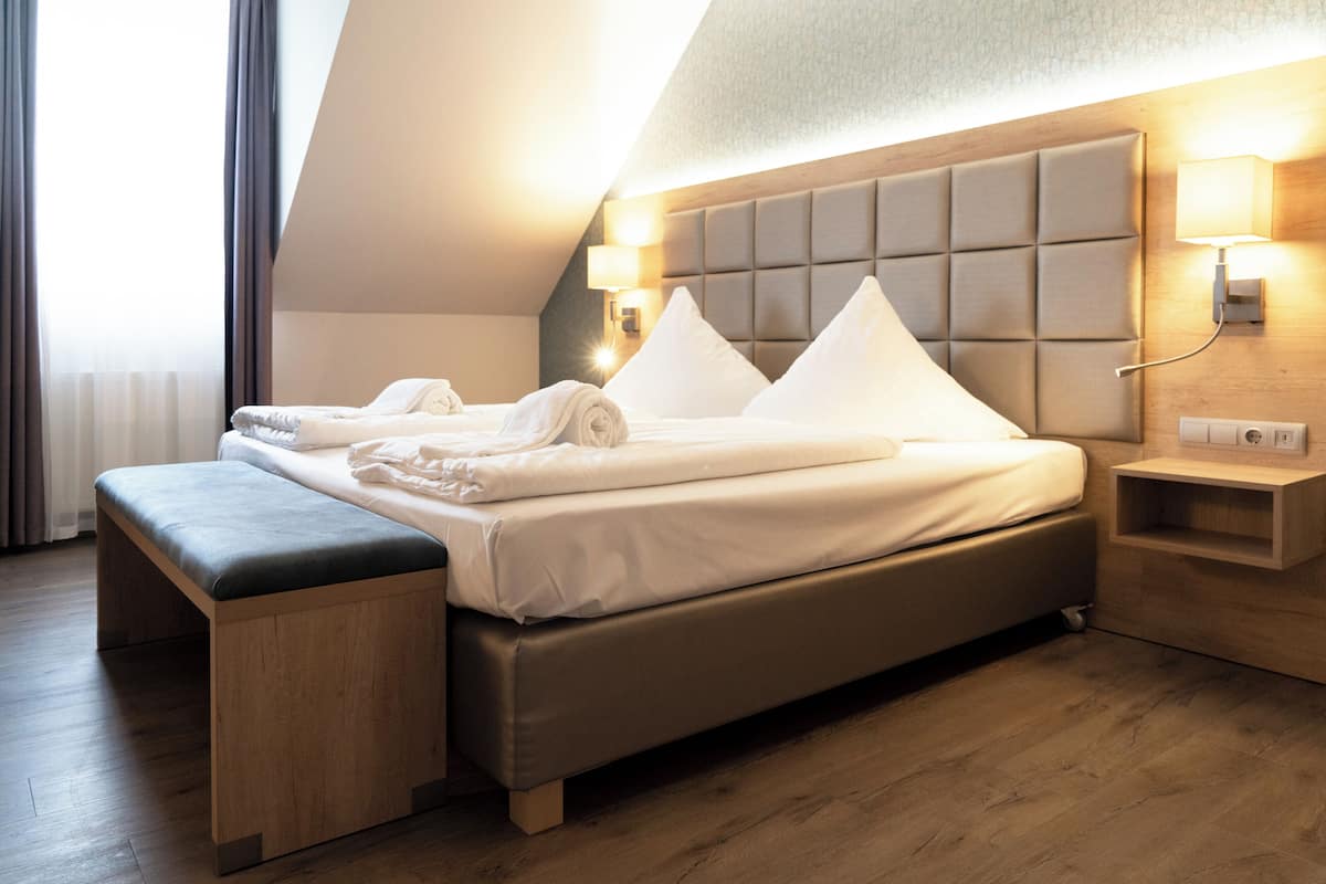 A modern hotel room with a large bed, a bench, a headboard with lights, and a wooden nightstand.