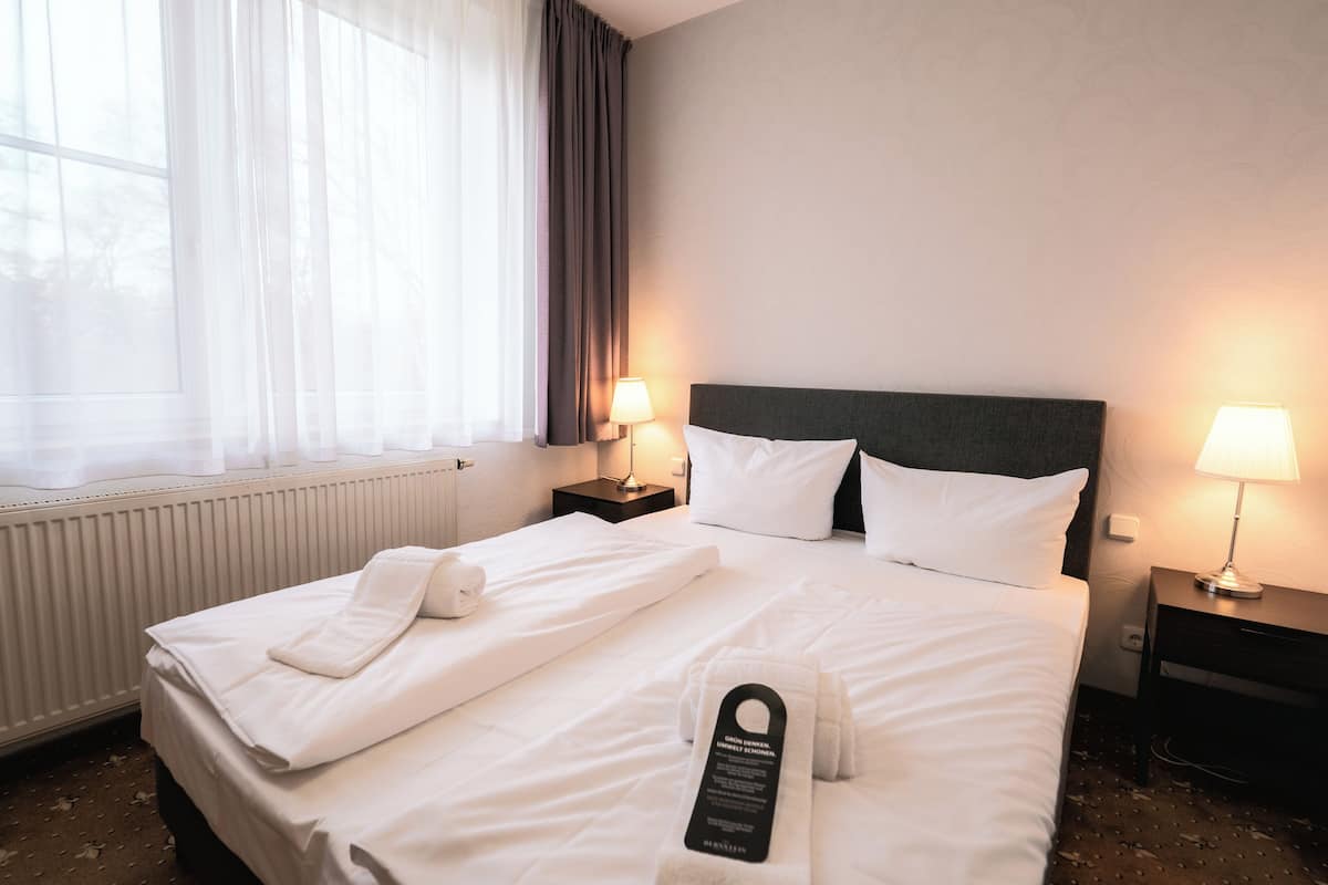 A hotel room with a neatly made bed, two bedside lamps, a window with curtains, and a radiator.