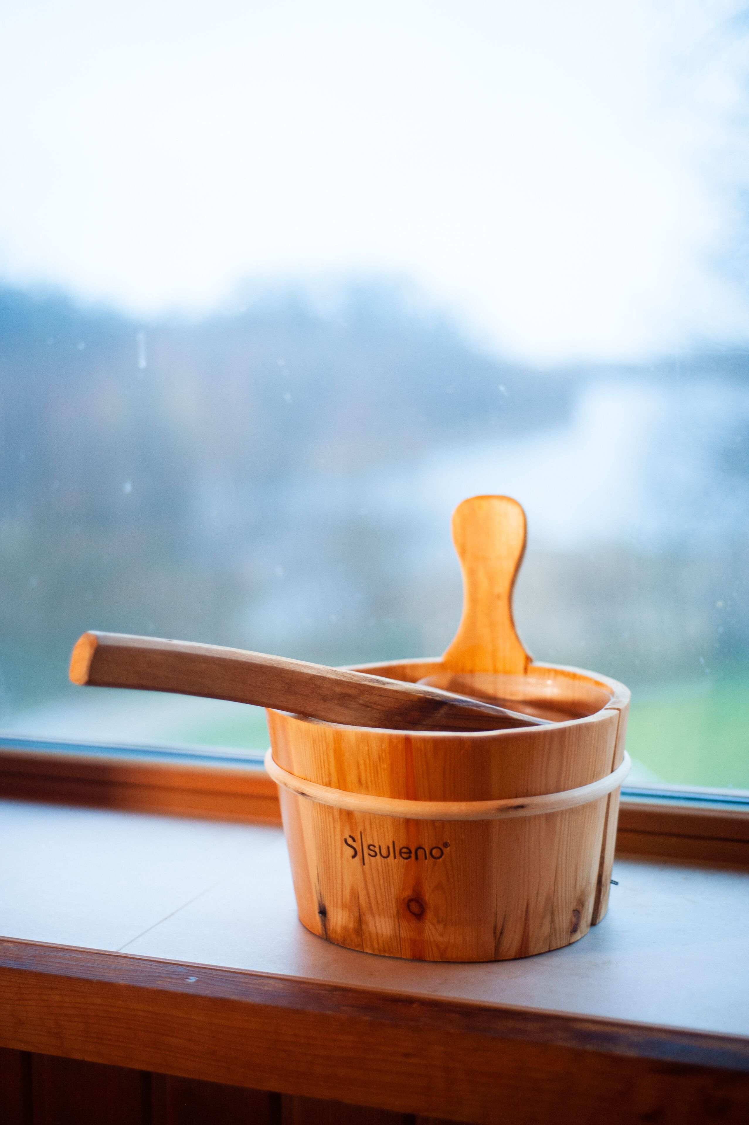Un cubo de madera con una cuchara de madera dentro, sobre un alféizar con una vista desenfocada de árboles y cielo al fondo.