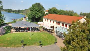 A riverside hotel with outdoor seating, a glass-enclosed dining area, and a red-roofed building.