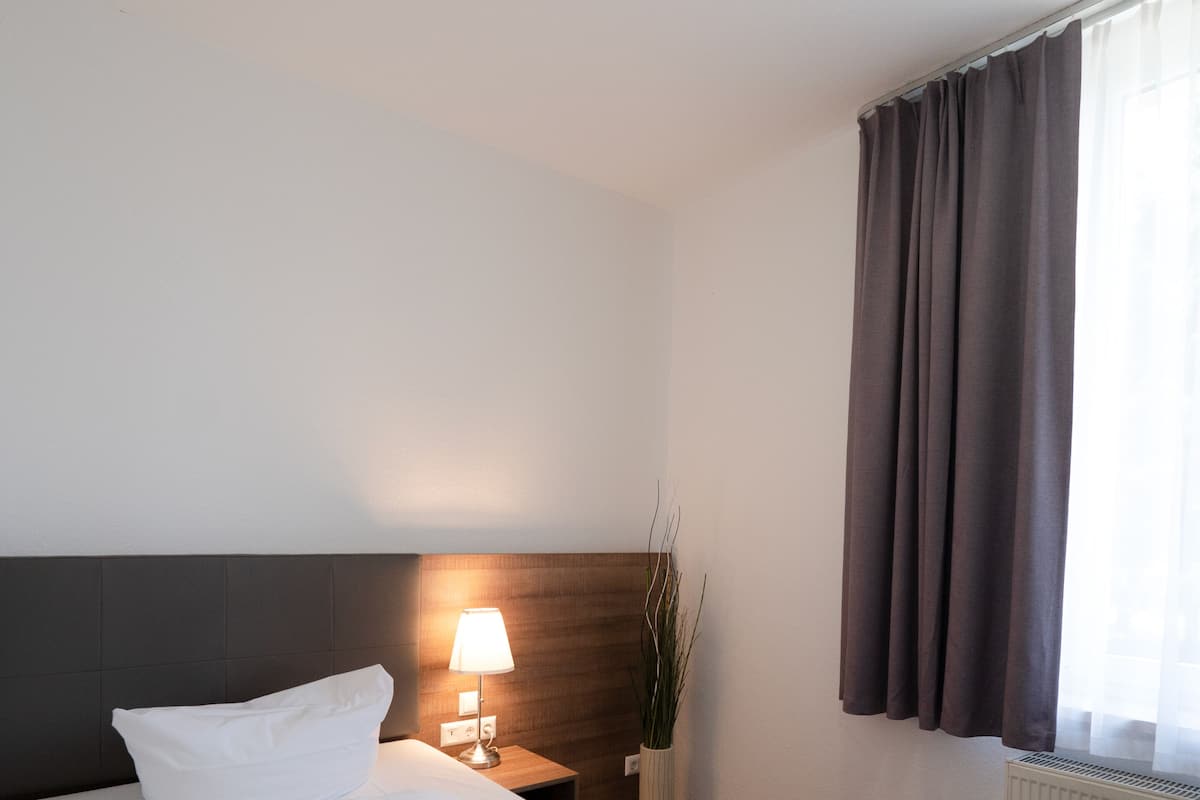 A neatly made bed with white linens, a bedside lamp, a potted plant, and a window with curtains.