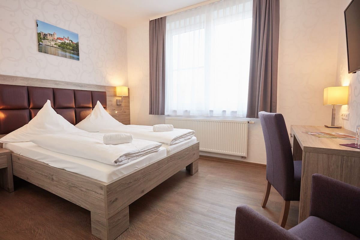 A hotel room with a bed, a desk, a chair, a window with curtains, a wall-mounted lamp, and a painting.