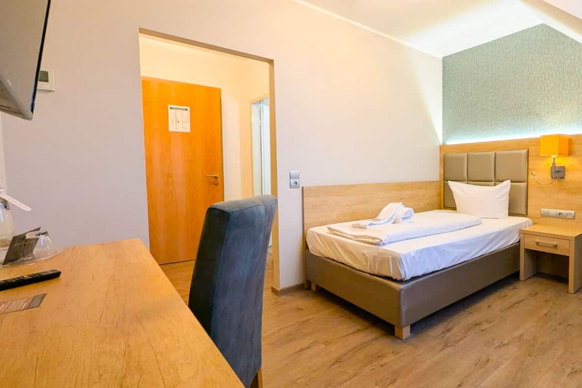 A hotel room with a bed, bedside table, chair, desk, and a door.
