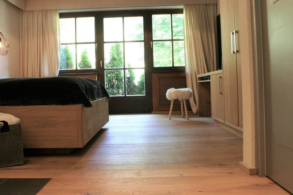 Ein Schlafzimmer mit großem Fenster, einem Bett, einem kleinen Hocker und einer Tasche auf dem Boden.