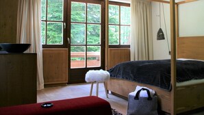 Un dormitorio con cama de madera, un taburete pequeño, un banco rojo y una ventana con cortinas.