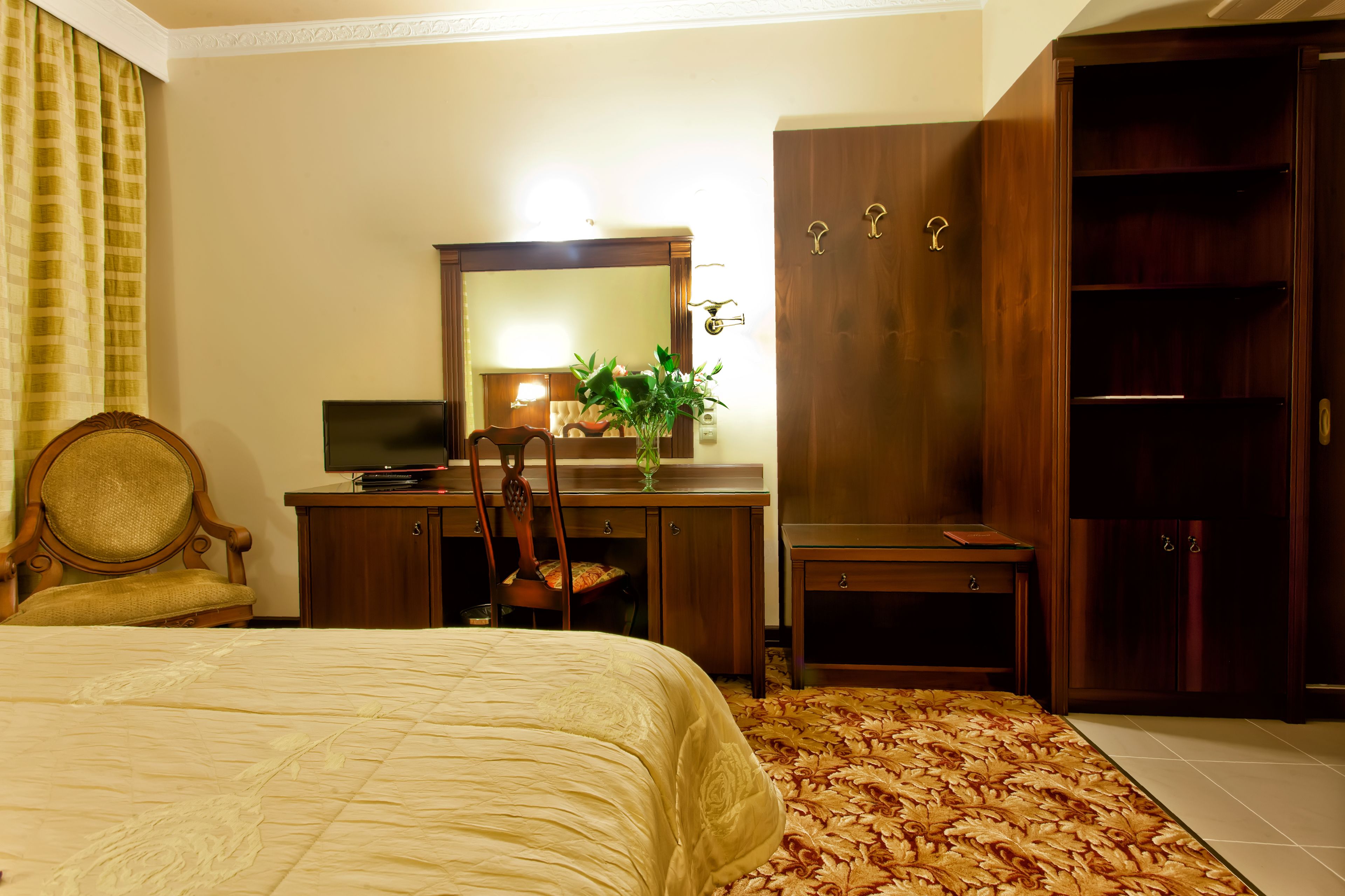 A hotel room with a bed, a desk with a mirror, a chair, a wardrobe, and a small table with a plant.