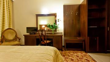 A hotel room with a bed, a desk with a mirror, a chair, a wardrobe, and a small table with a plant.