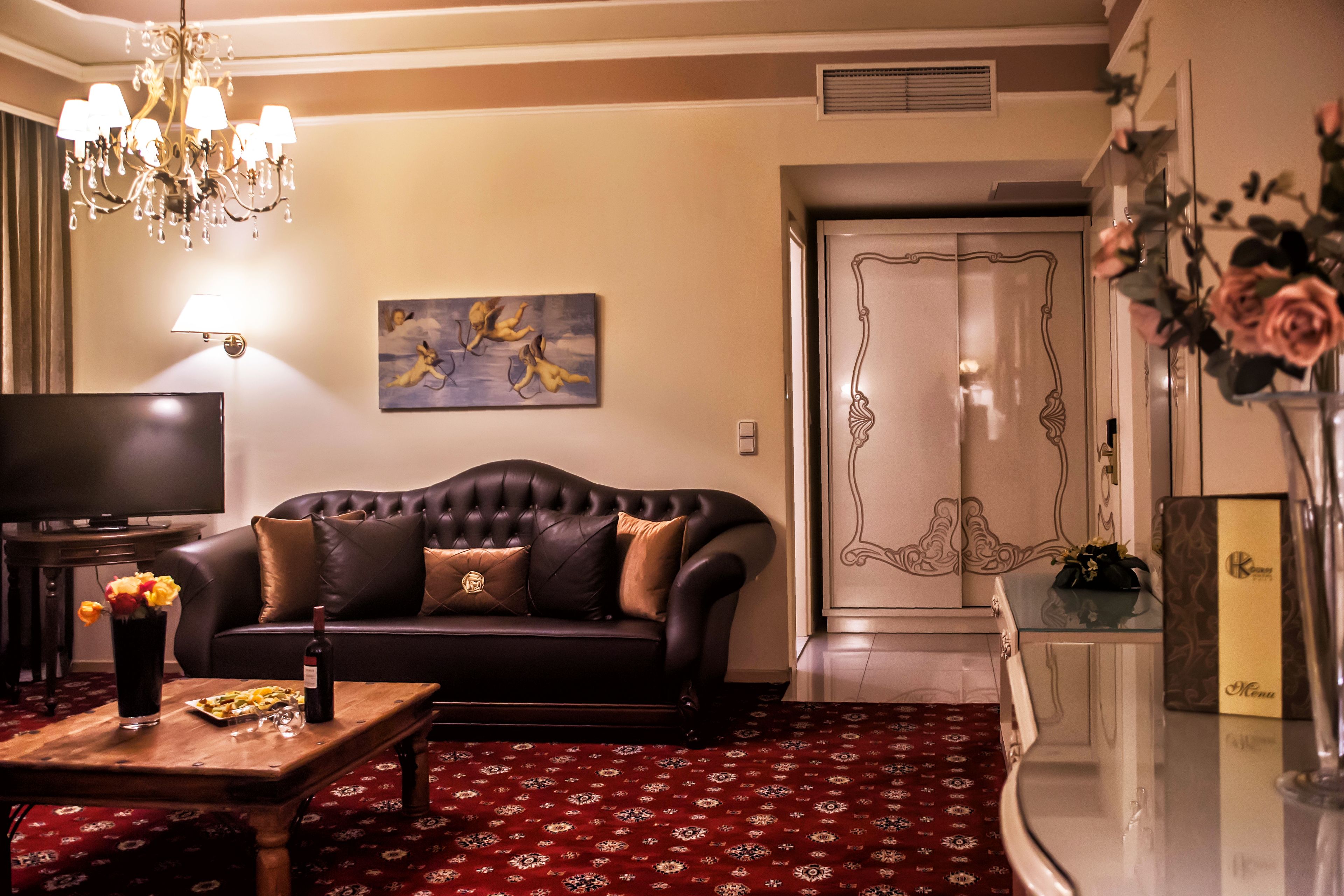 A living room with a dark leather sofa, a wooden coffee table with a bottle and flowers, a flat-screen TV, a chandelier, and a red patterned carpet.