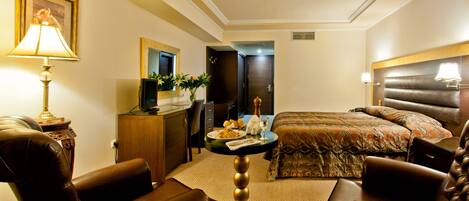 A hotel room with a bed, two leather armchairs, a desk with a lamp, a television, and a dining table with a vase of flowers.