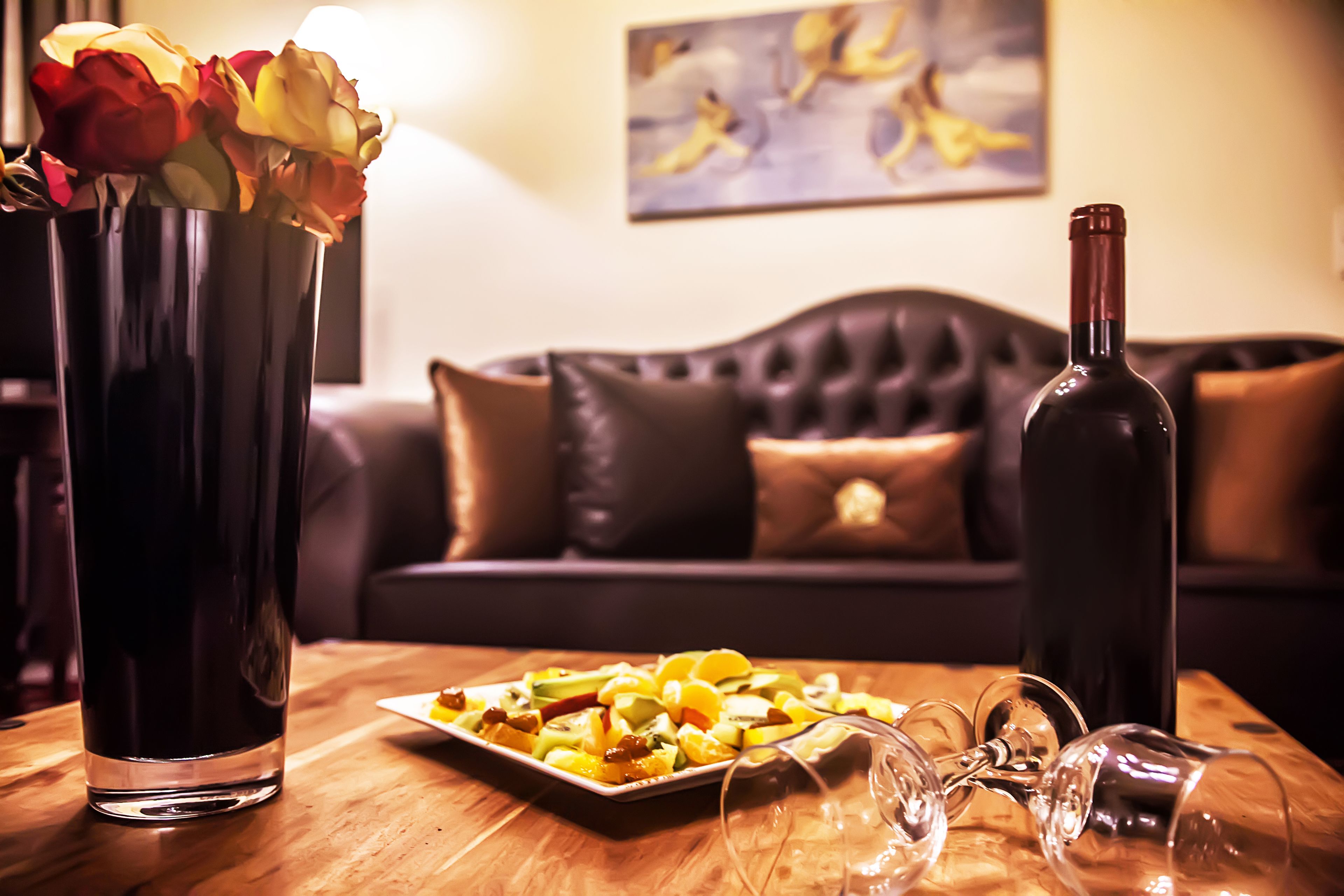 Une table à manger avec une assiette de fruits, une bouteille de vin et un vase de fleurs.