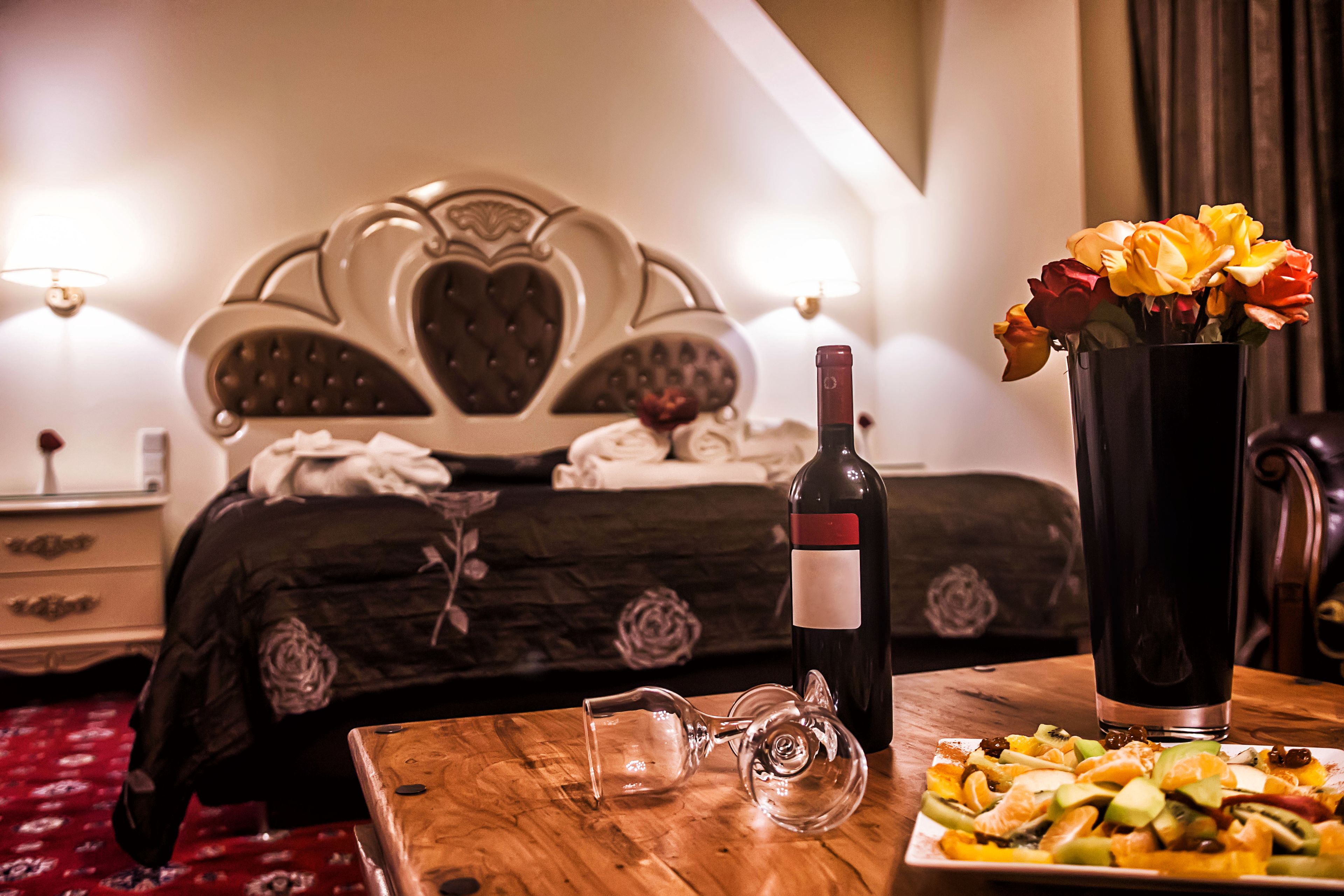 A hotel room with a bed, a wooden table with fruit, a bottle of wine, and a vase with flowers.