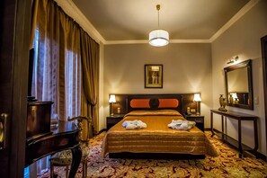 Una habitación de hotel con una cama grande, dos mesitas de noche con lámparas, un espejo, un cuadro en la pared y un piano en la esquina.