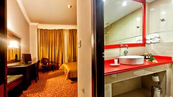 Una habitación de hotel con cama, escritorio con computadora, silla, baño con lavamanos de color rojo y espejo.