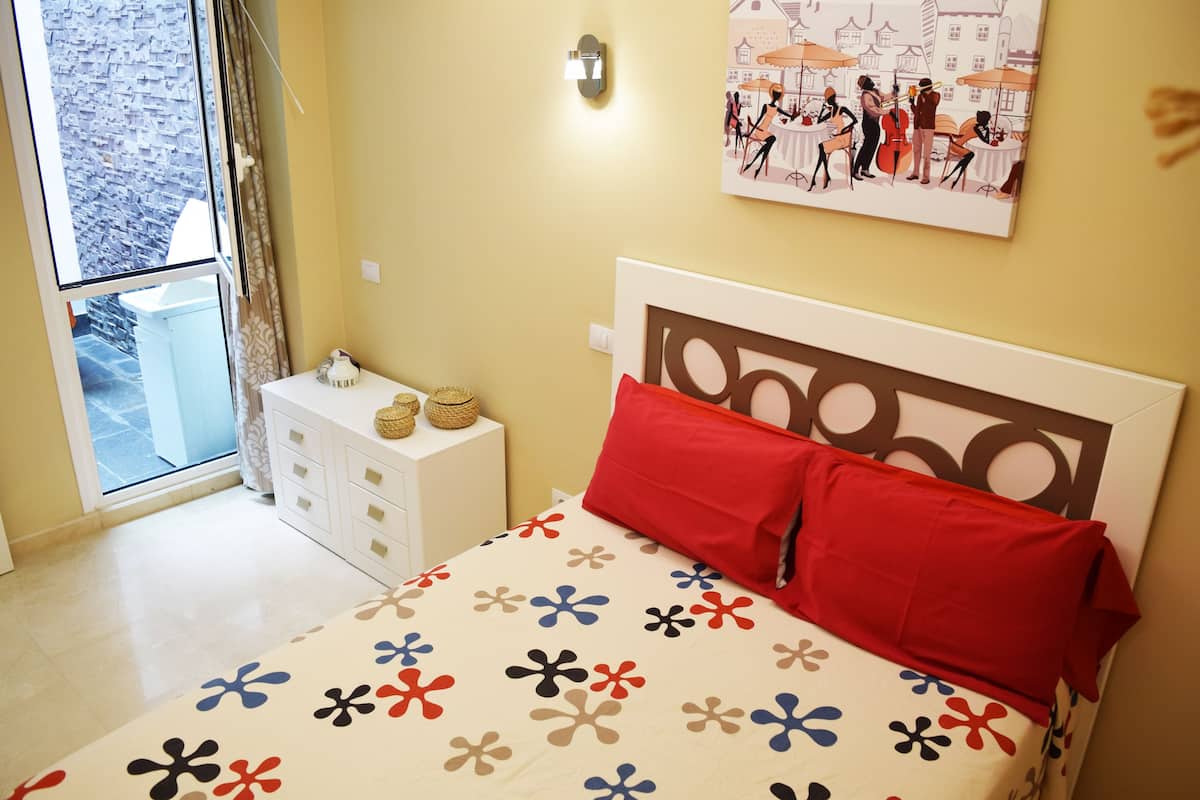 Un dormitorio con cama, mesita de noche, ventana y lámpara fijada a la pared.