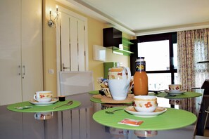 A dining area with a table set for two, a pitcher, and a bottle of juice. There are two chairs and a window with curtains.