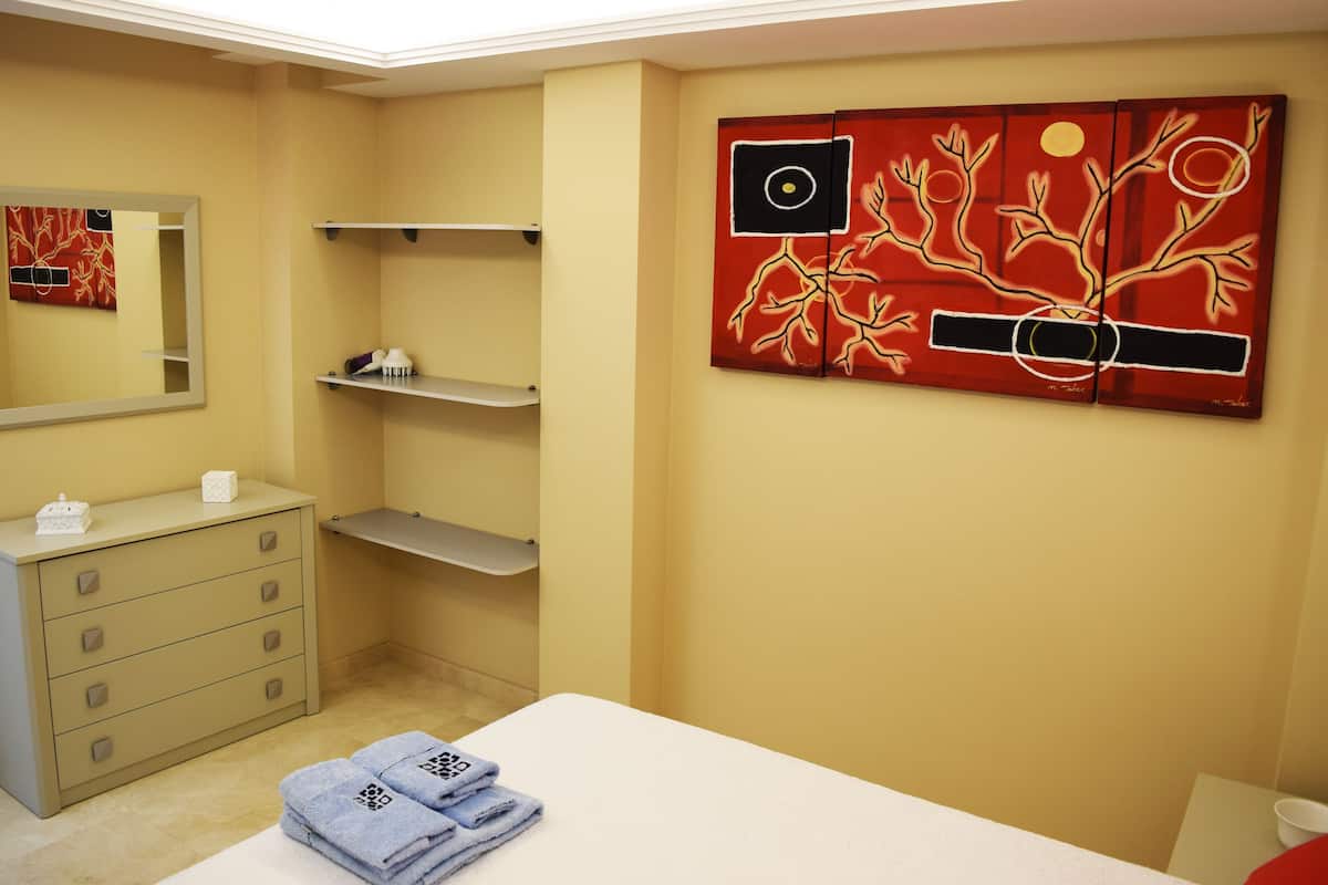 Habitación de hotel con cama, cómoda, espejo, una obra de arte en la pared y repisas con artículos de aseo.