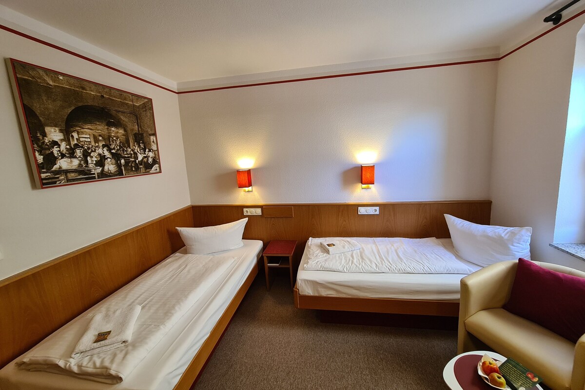 Une chambre d’hôtel avec deux lits simples, une petite table sur laquelle se trouvent un livre et des fruits, un canapé et un tableau accroché au mur.