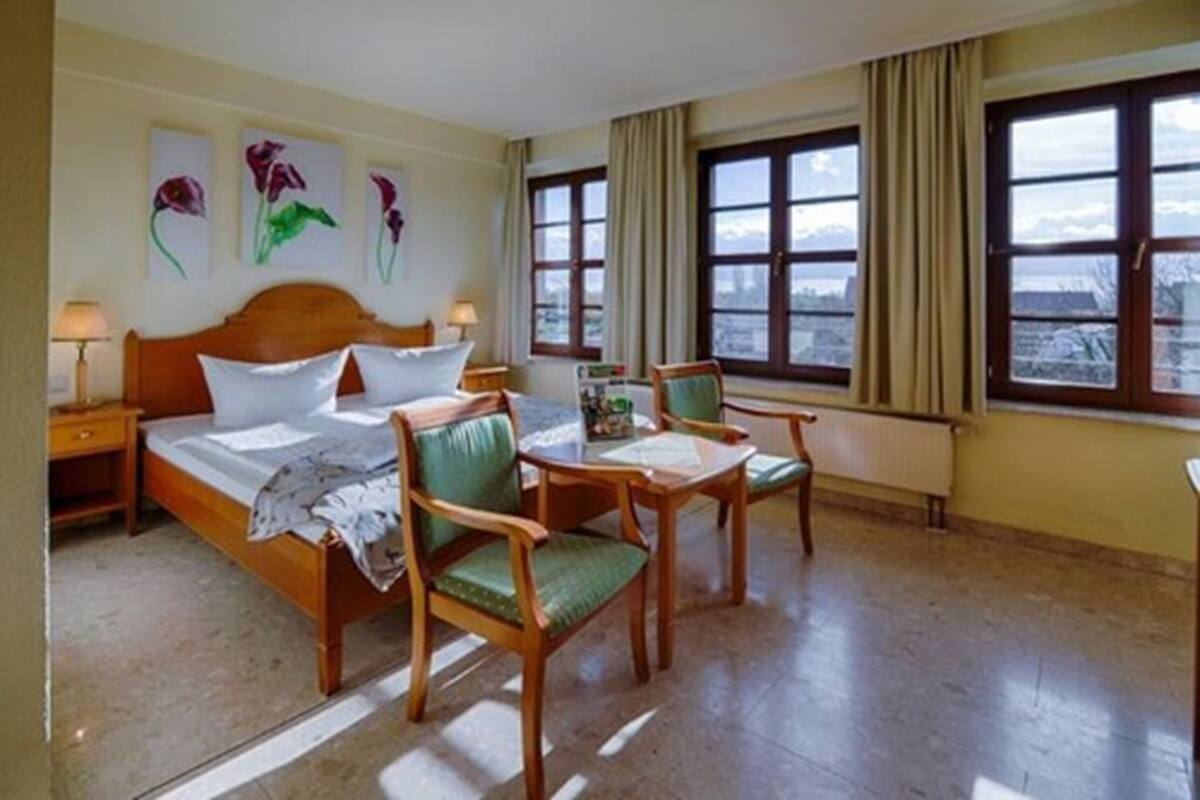 A hotel room with a bed, two chairs, a table, and a view of the city through large windows.
