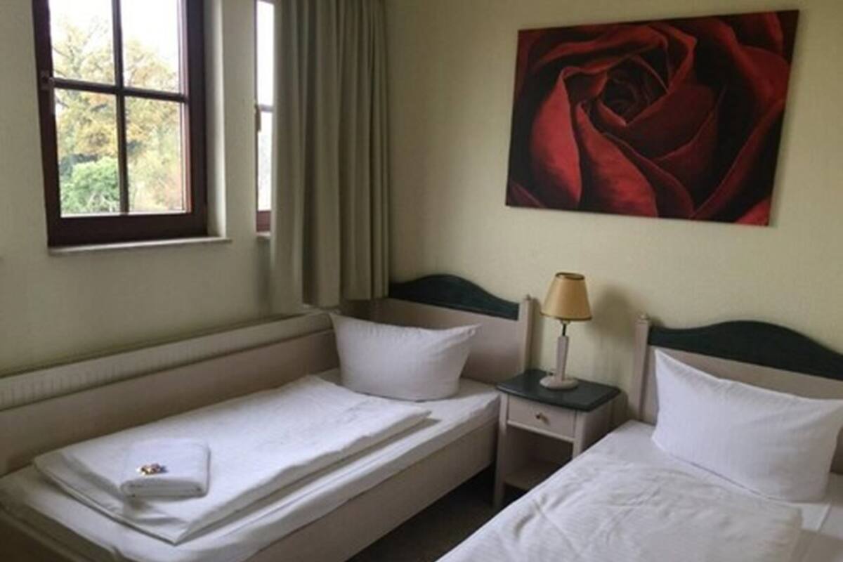 A hotel room with two single beds, a nightstand with a lamp, and a large window with curtains.