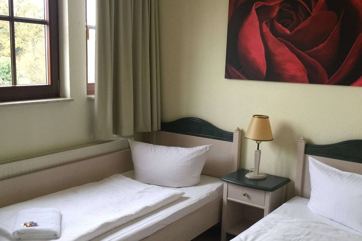A hotel room with two single beds, a nightstand with a lamp, a window with curtains, and a painting of a rose on the wall.