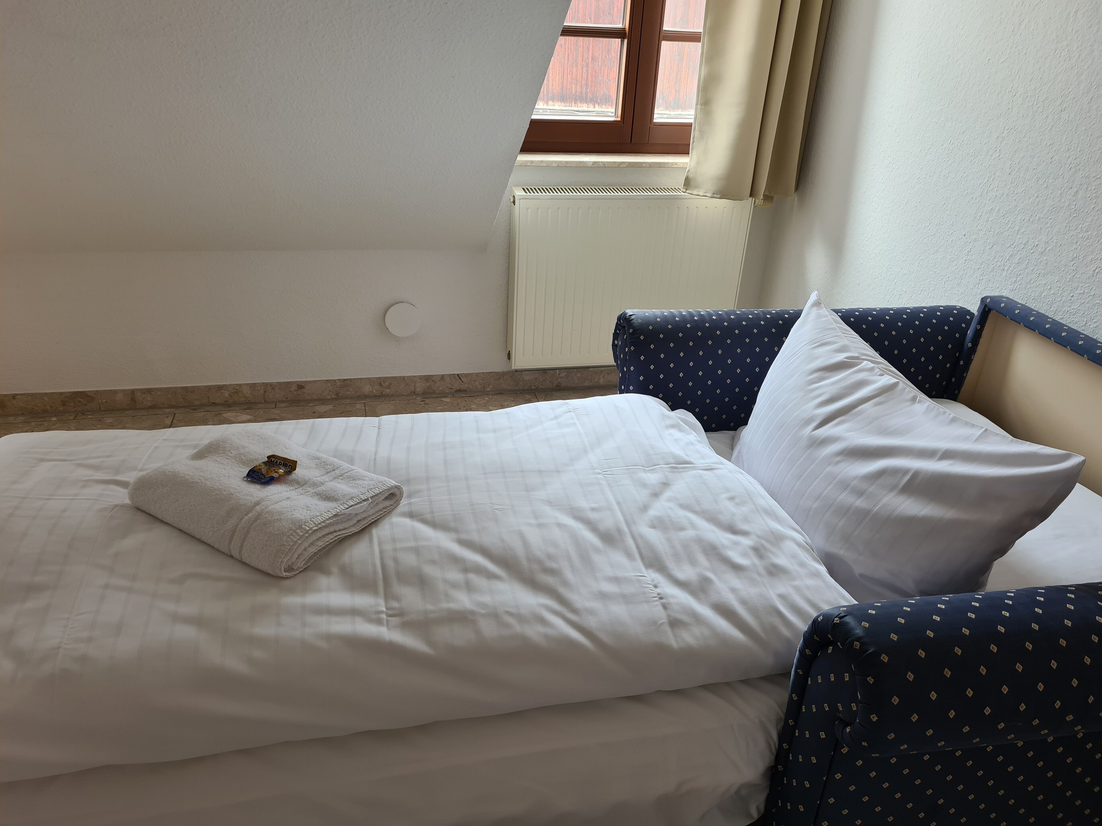 A neatly made bed with white linens and a folded towel, a window with curtains, and a wall-mounted radiator.