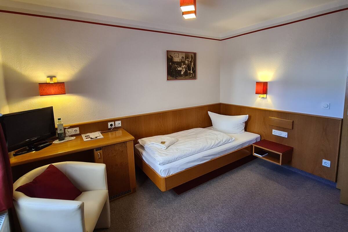 A hotel room with a bed, a desk, a chair, a television, and two wall-mounted lamps.