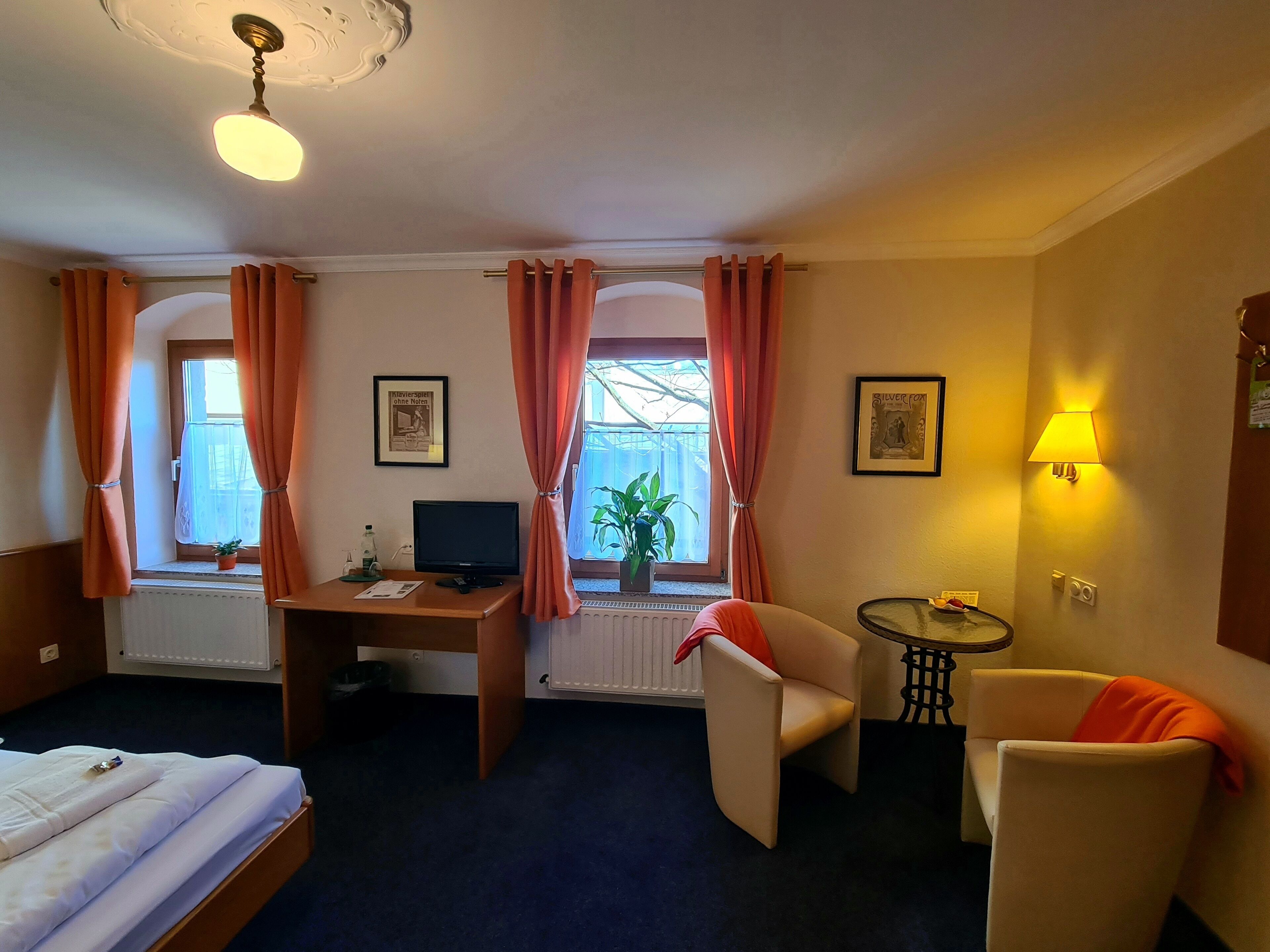 Una habitación de hotel con una cama, un escritorio con computadora, dos sillas, una mesita, una lámpara y un ventanal con cortinas.