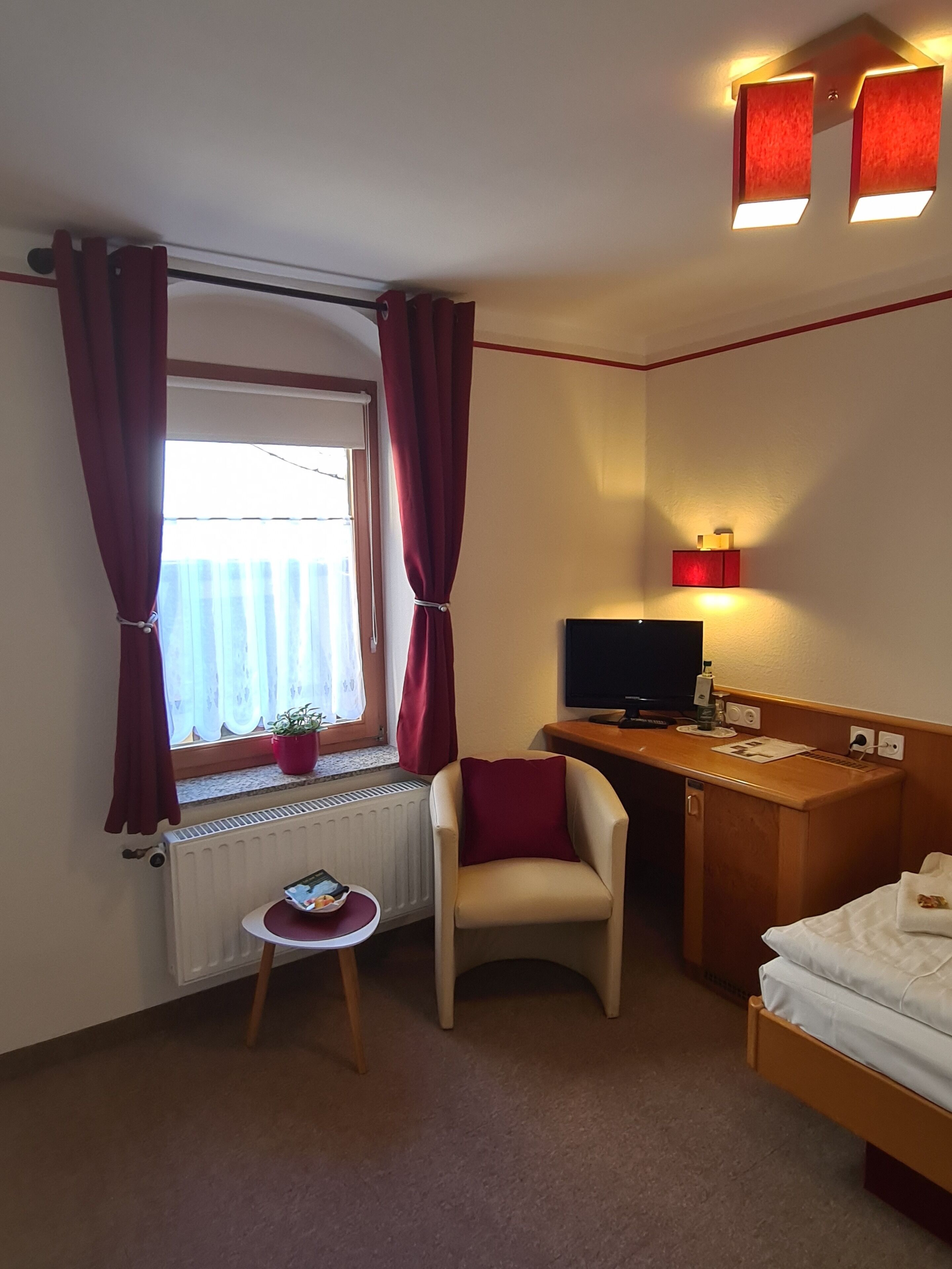 Habitación de hotel con cama, escritorio con computadora, silla, ventana con cortinas, radiador y televisor montado en la pared.