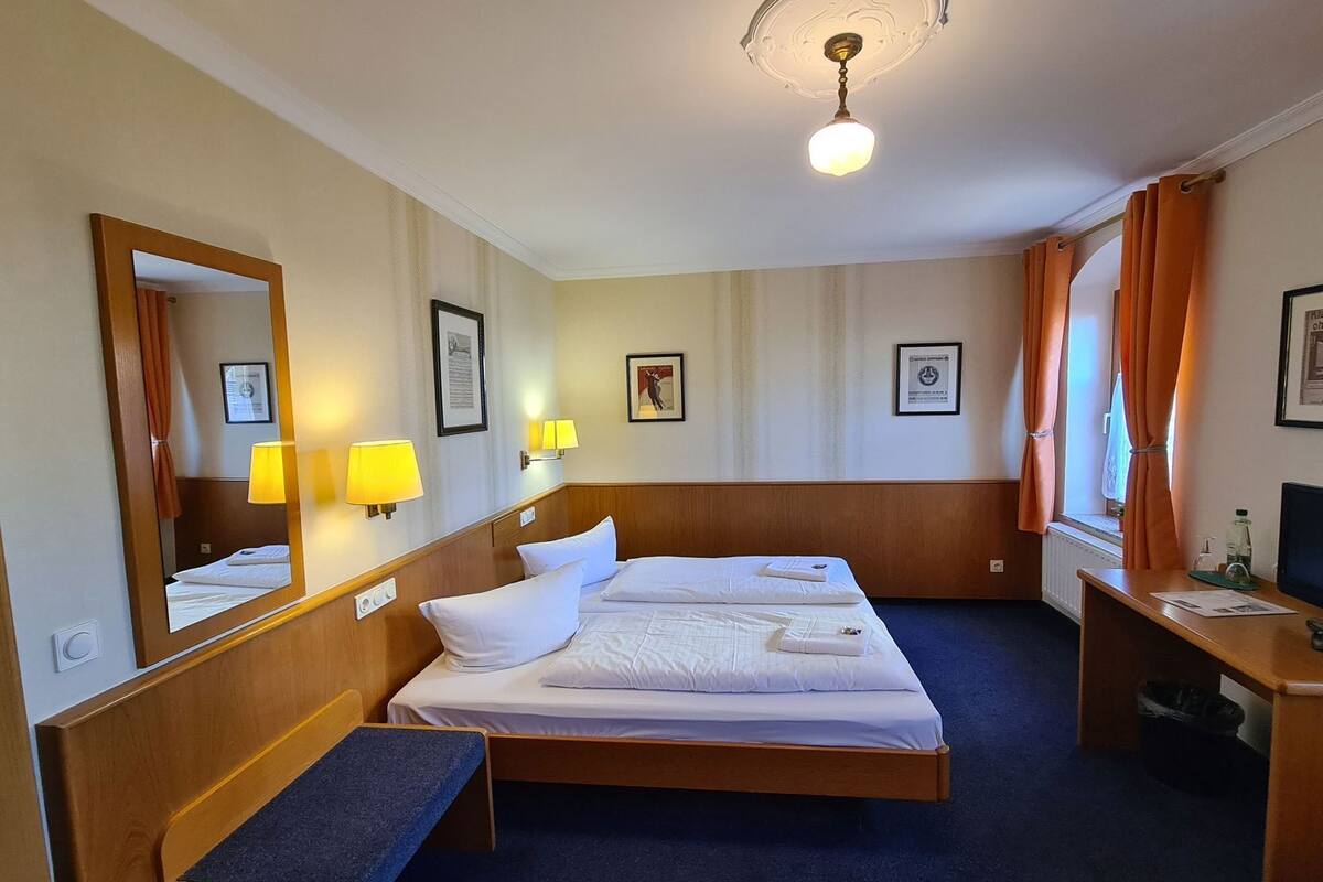 A hotel room with two beds, a desk, a chair, a bench, a ceiling light, and a window with curtains.
