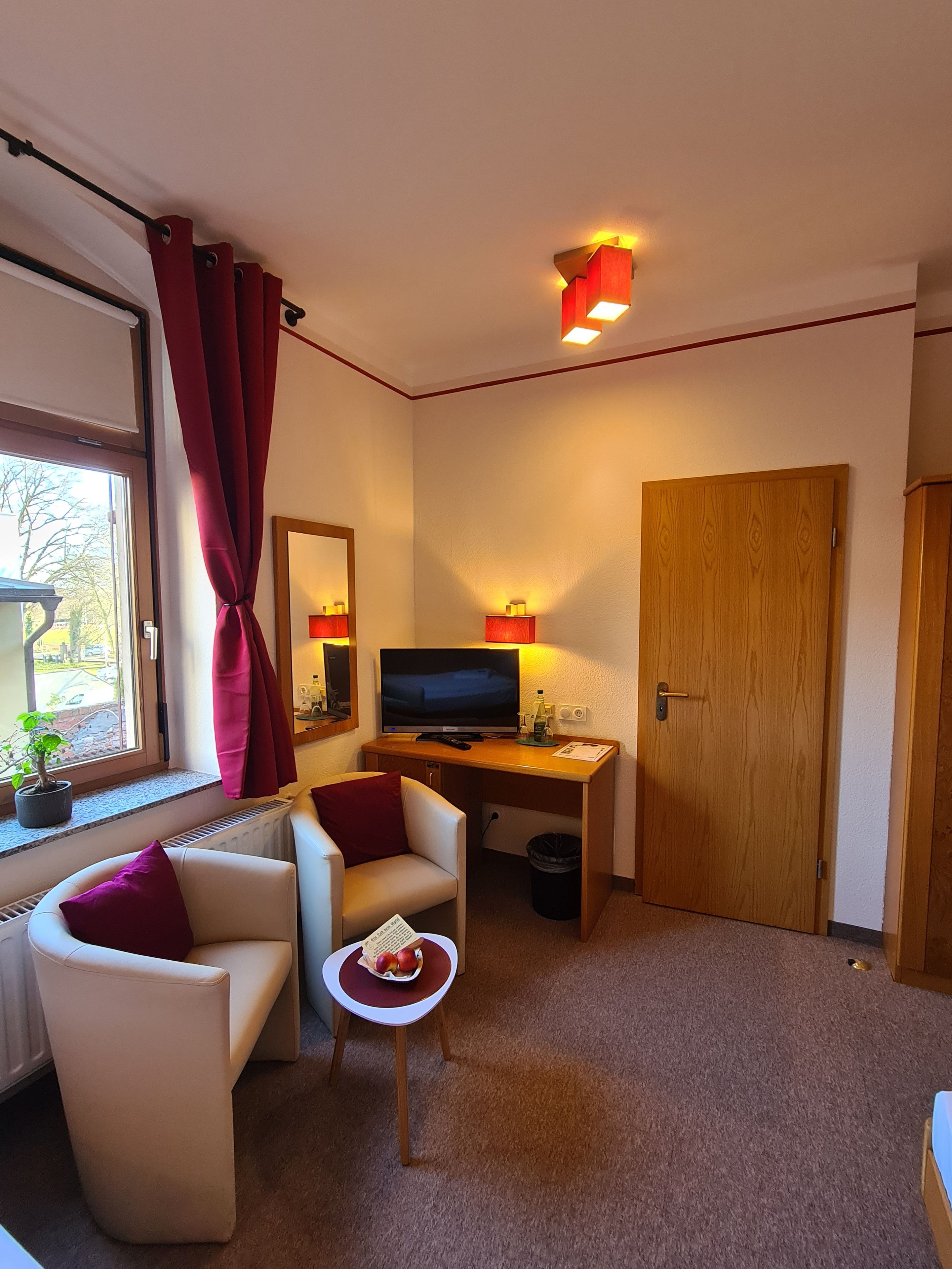 A compact room with a television, a desk, a chair, a small table with a bowl of fruit, and a window with curtains.