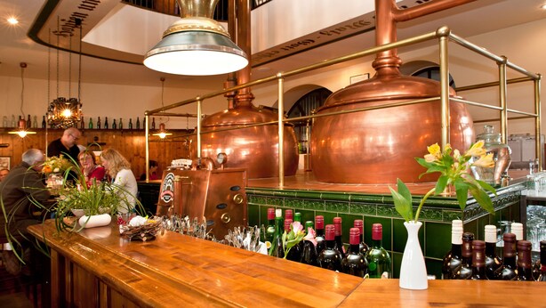 Een bar met koperen brouwapparatuur, een houten toog en flessen wijn.
