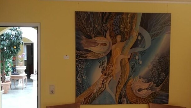 Una habitación con un cuadro abstracto grande en la pared, dos sillas de madera con cojines negros, una mesita de madera pequeña y un sofá rojo y naranja con estampado.