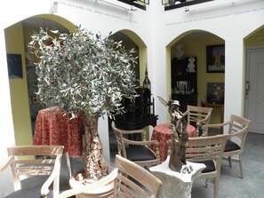 Une cour ensoleillée avec un grand arbre, des chaises en bois et une petite table agrémentée d’objets décoratifs.