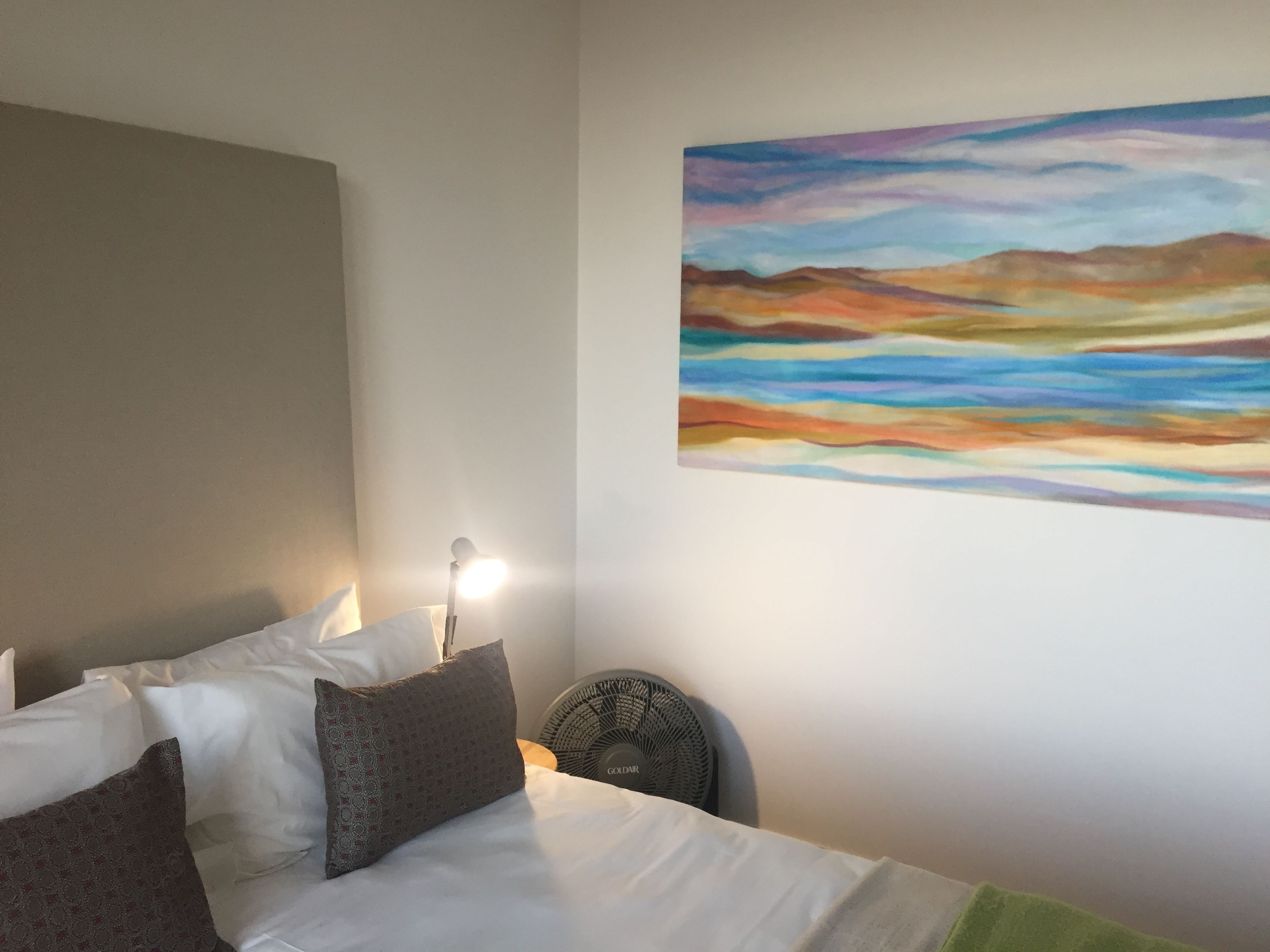 A hotel room with a bed, pillows, a wall-mounted painting, and a standing fan.