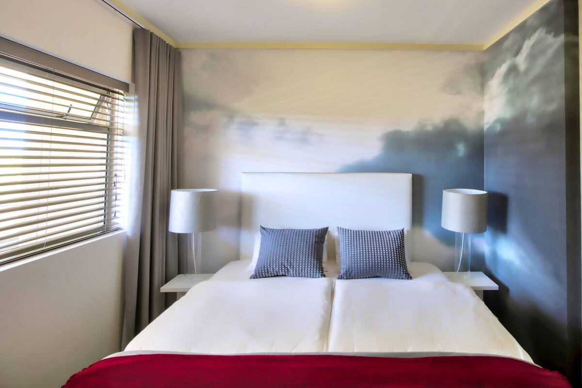 A hotel room with a large bed, two bedside lamps, a red throw blanket, and a wall mural.
