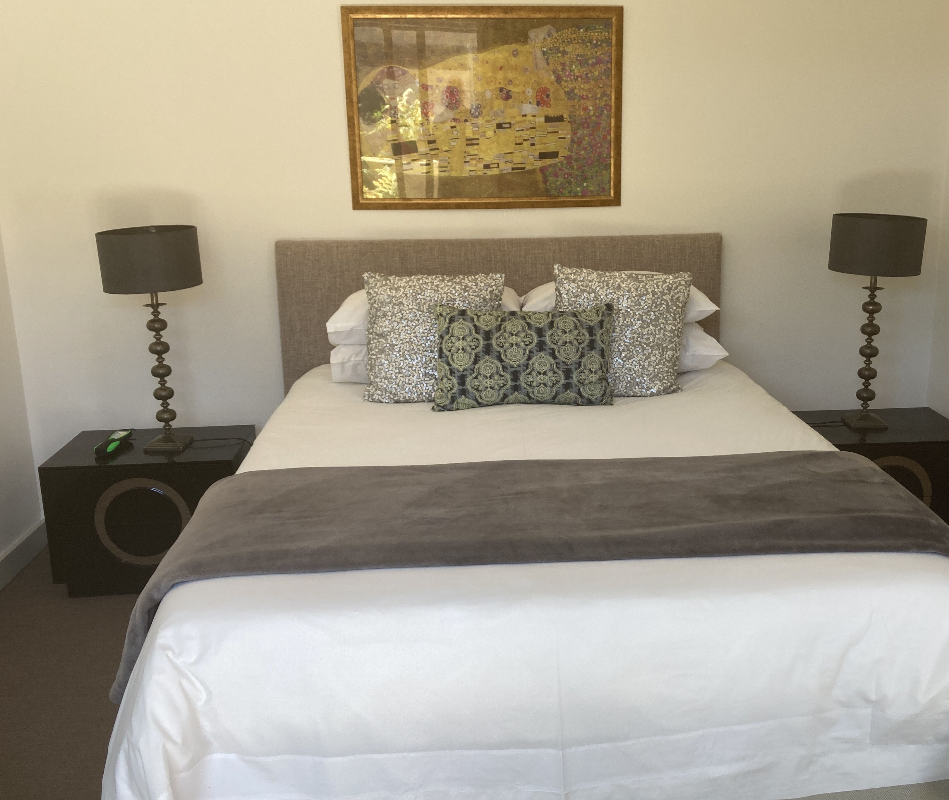 A neatly made bed with a white comforter and patterned pillows, flanked by two bedside tables with lamps, and a framed artwork above the bed.