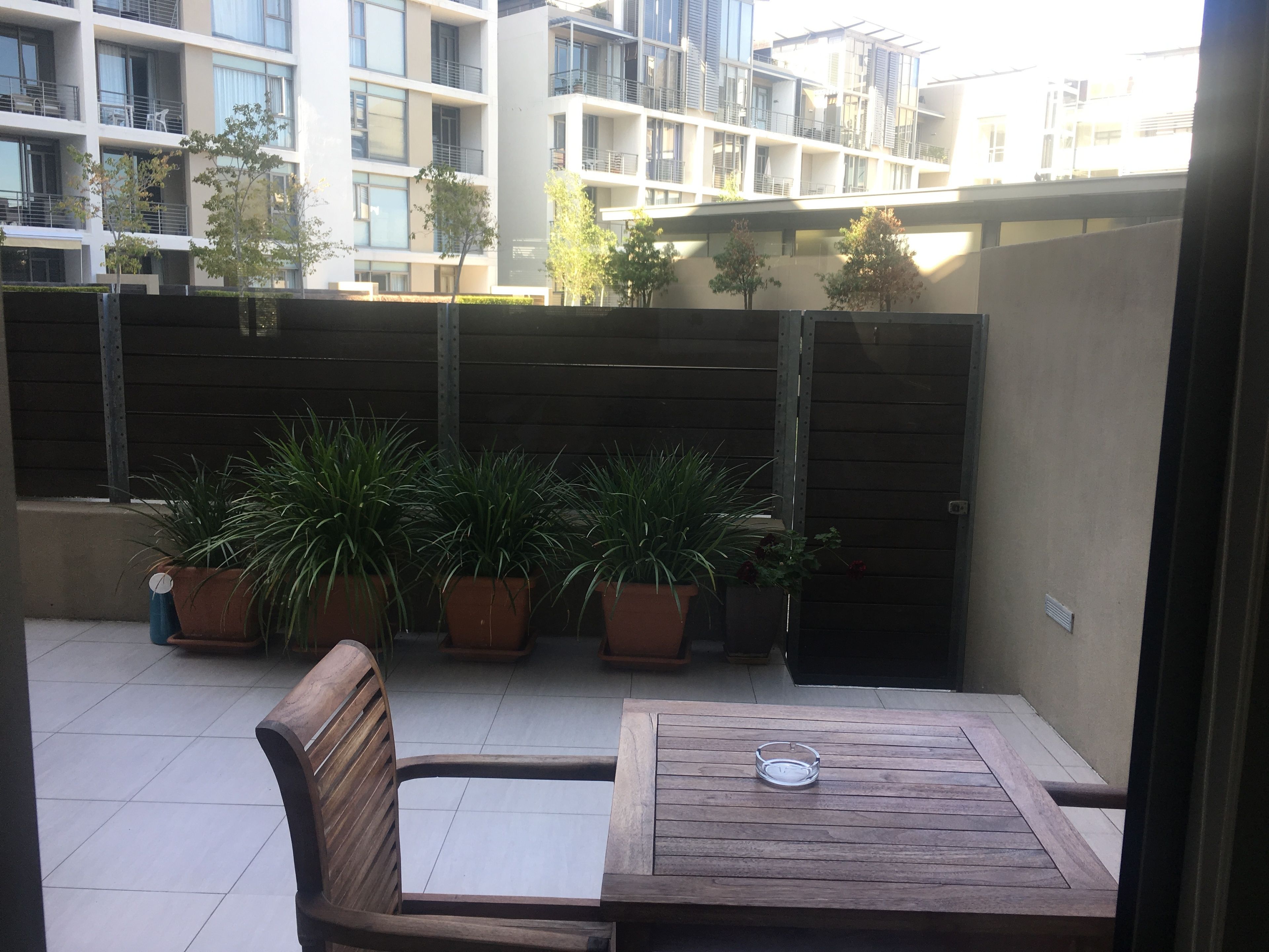 A wooden table and bench in an outdoor patio area with potted plants and a view of a modern building in the background.