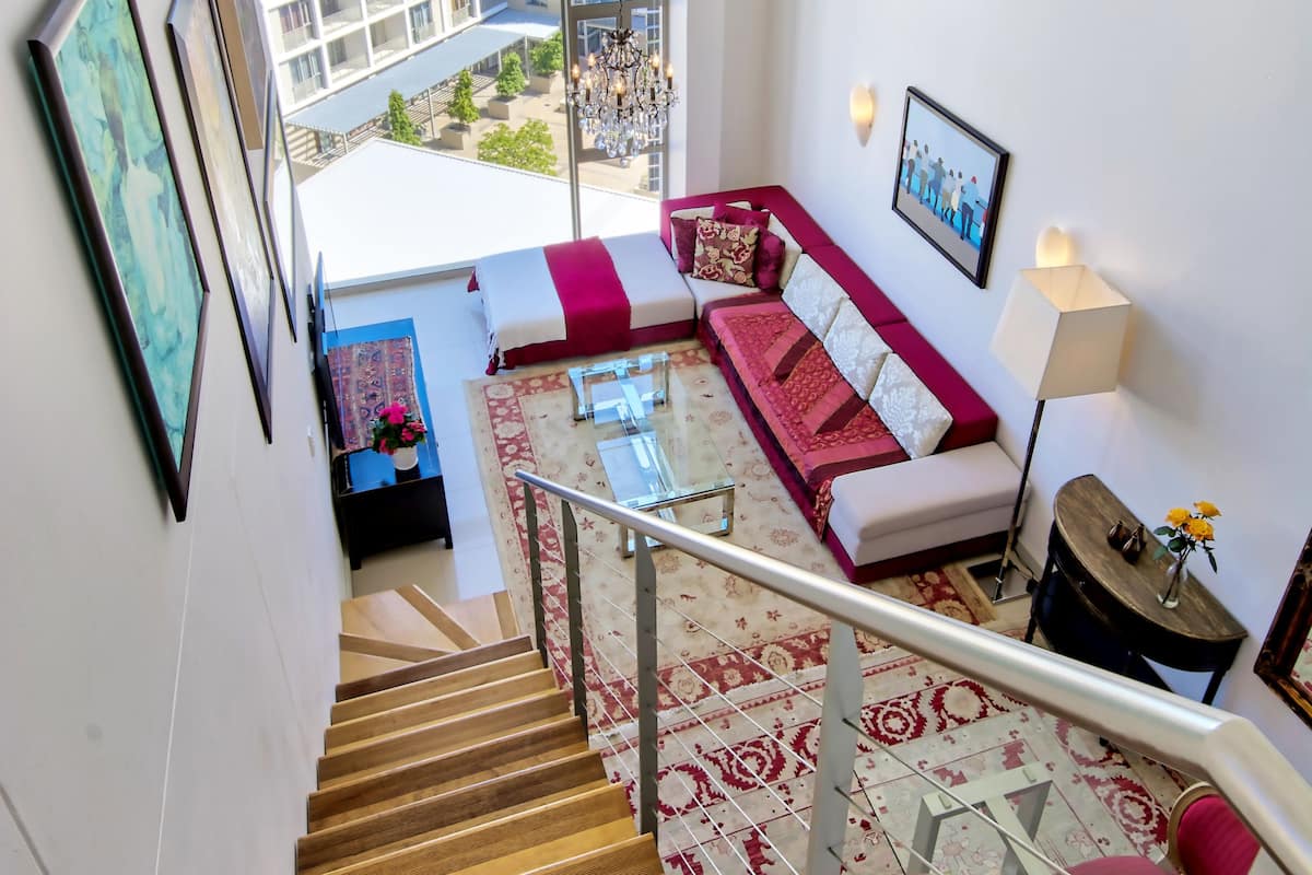 A modern living area with a staircase, a red and white sofa, a glass coffee table, and a view of buildings outside.