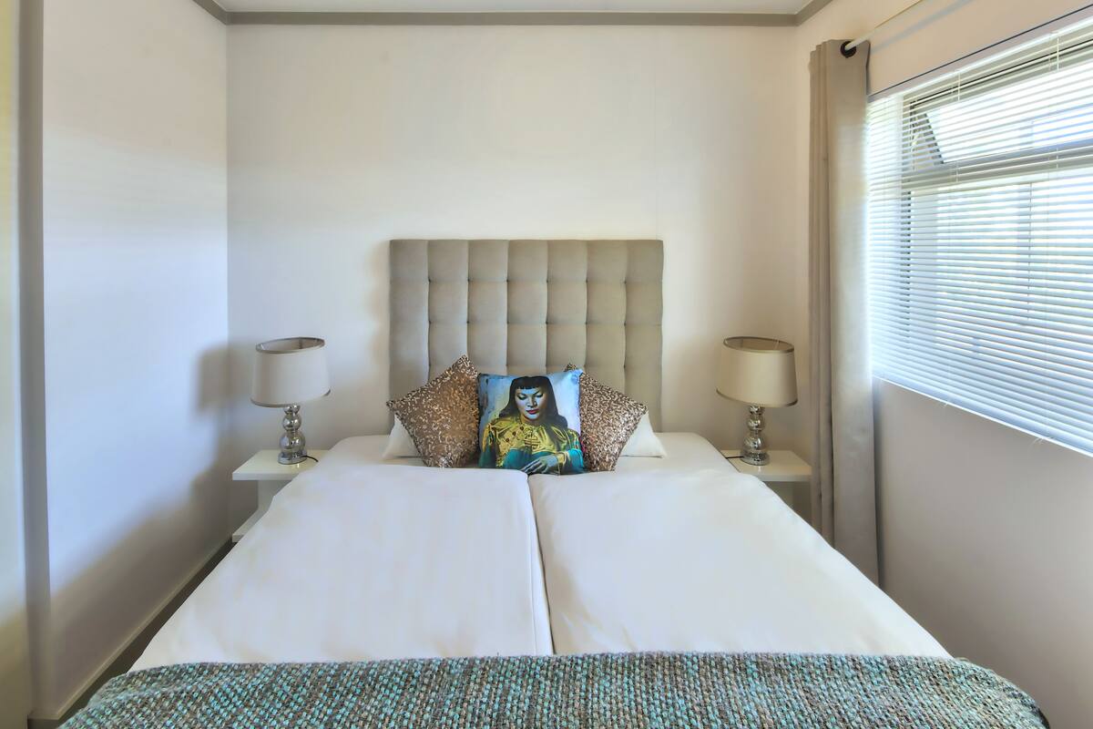 A neatly made bed with a tufted headboard, two bedside lamps, and a window with blinds.