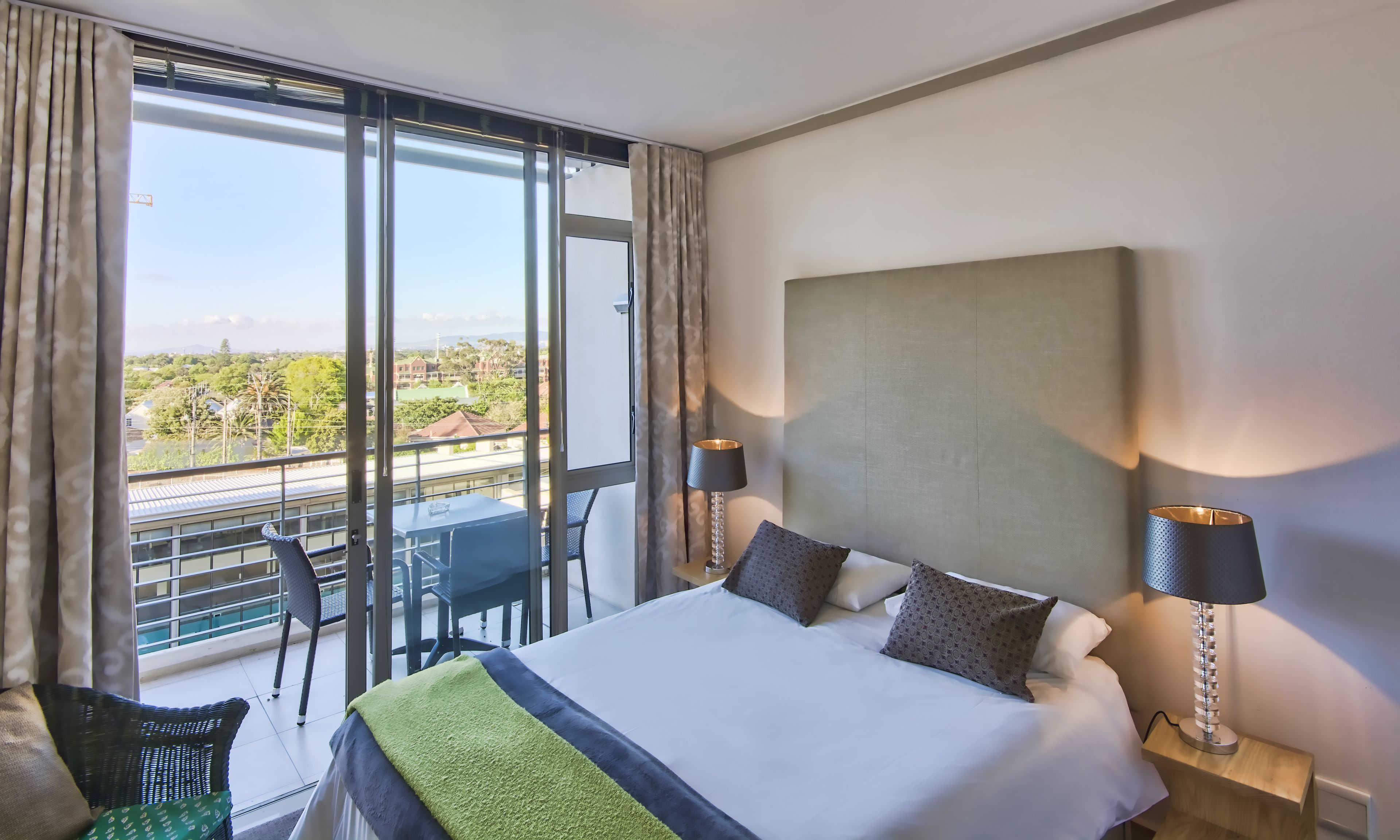 A hotel room with a large bed, two bedside lamps, a view of the cityscape, and a balcony with a table and chairs.