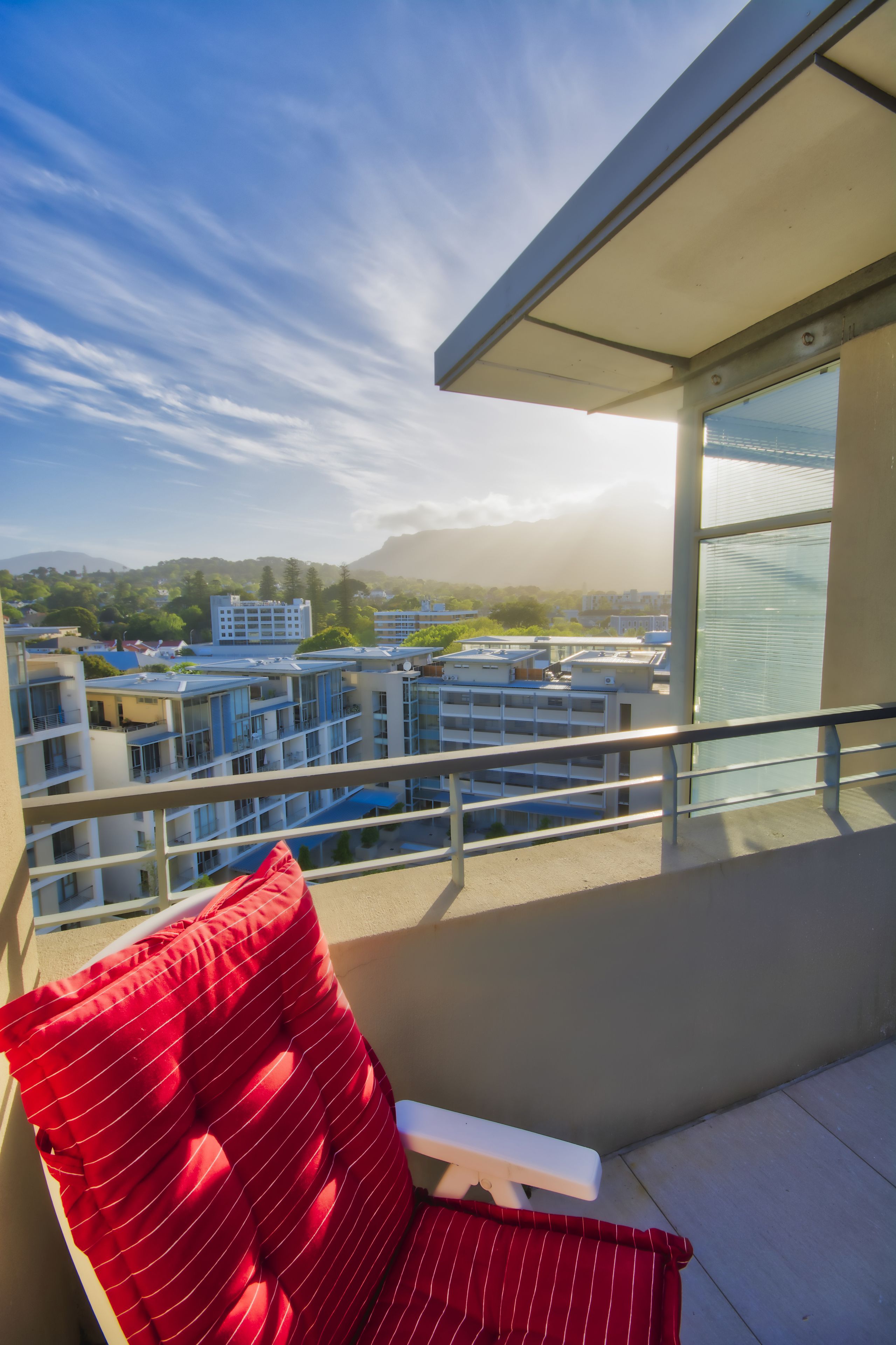 A balcony with a red-striped cushion and a white chair, overlooking a cityscape with modern buildings and a clear sky.