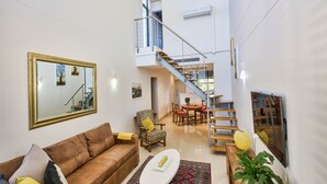 En rummelig opholdsstue med en lædersofa i brunt, et rundt sofabord og et tæppe med et mønster. En trappe fører op til en etage med en balkon.