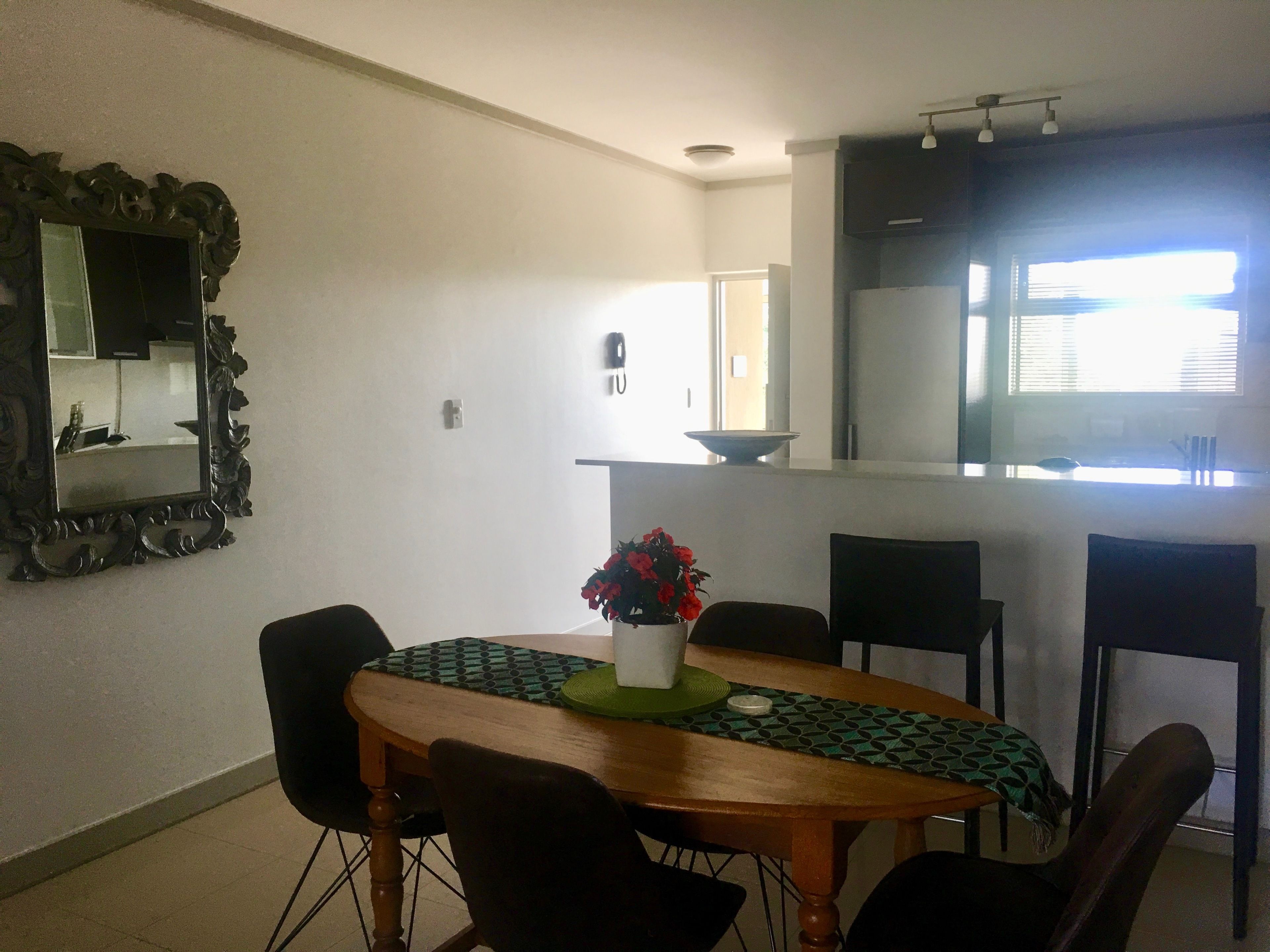A dining area with a wooden table, chairs, and a vase with flowers. There is a mirror on the wall and a kitchen area in the background.