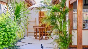 Un área de estar exterior de madera con mesa y sillas, rodeada de plantas tropicales y una estructura con techo de paja.