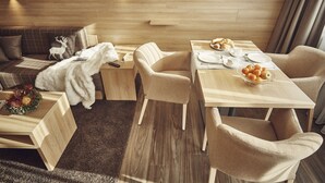 A modern hotel room with a wooden floor, a sofa with a reindeer pillow, a dining table set with plates, a bowl of fruit, and a white robe on the bed. - Private Holiday Homes by SOLARIA (Davos)