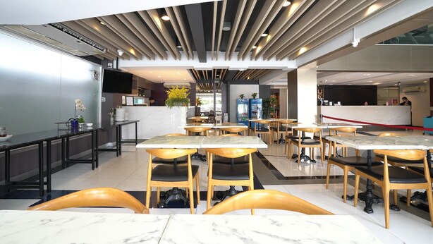 A modern restaurant interior with marble-topped tables and wooden chairs, a kitchen area in the background, and a ceiling with linear lighting fixtures. - Cubic Bed Pratunam - Hostel (Bangkok)