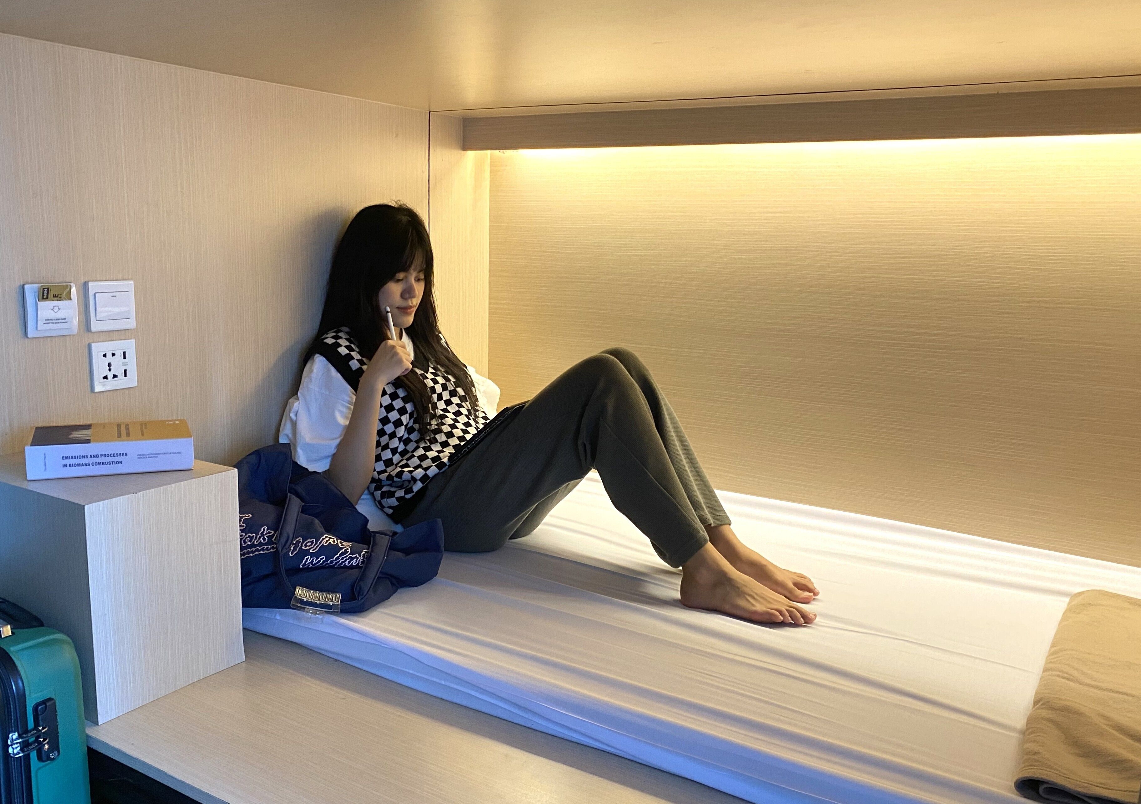 A single bed in a hotel room with a checkered top, a suitcase, and a box labeled "EMERGENCY AND PROCESSING IN CASE OF FIRE".