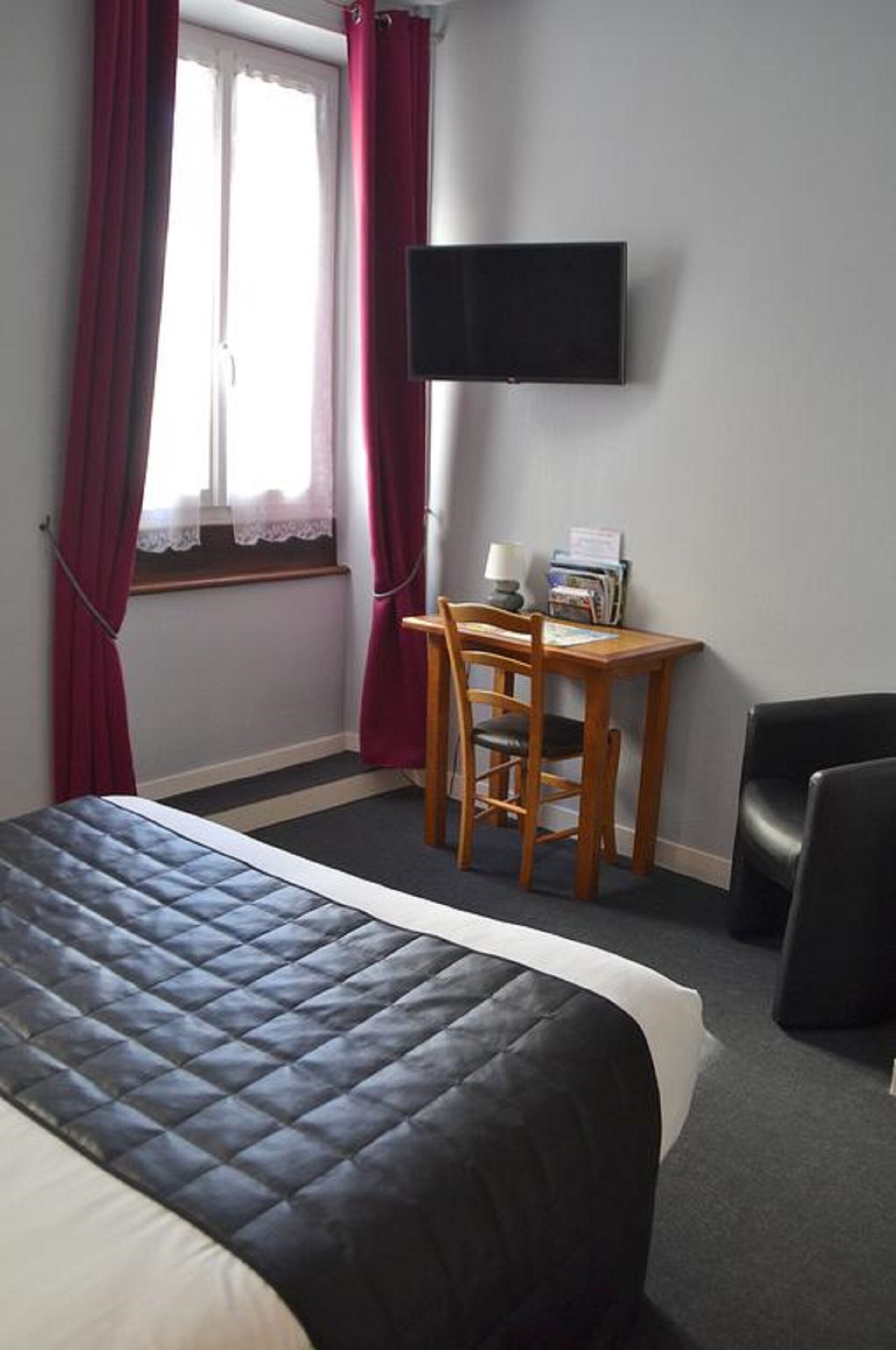 A hotel room with a bed, a desk with a chair, a television mounted on the wall, and a window with curtains.