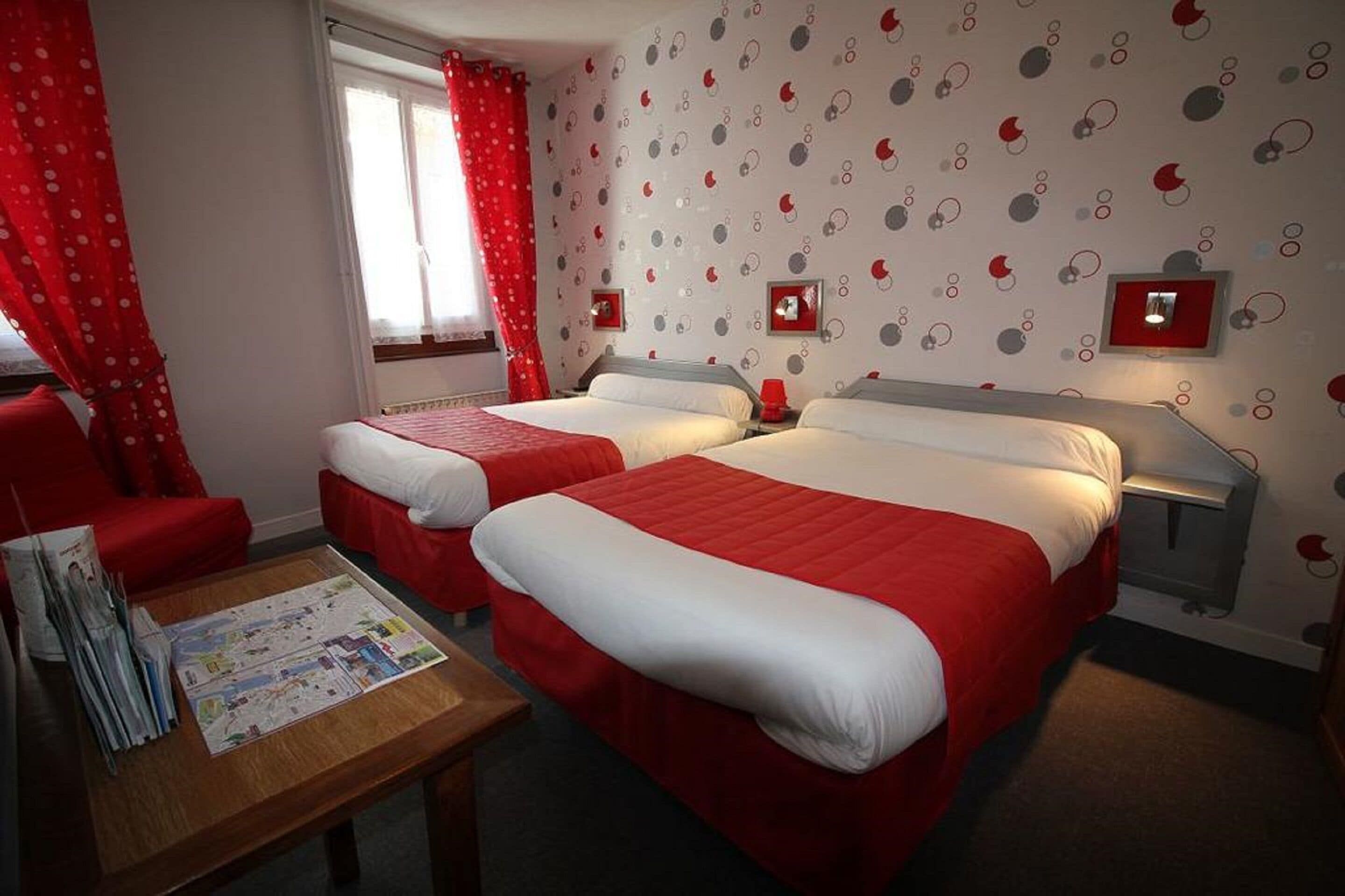 Habitación de hotel con dos camas, una mesa de madera con revistas, cortinas rojas y una pared con diseño circular.